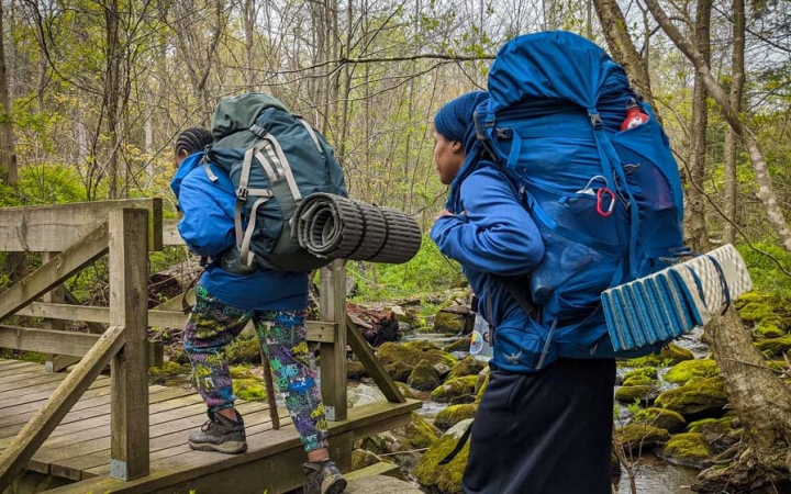 two young people wearing backpacks cross a creek over a bridge on a backpacking trip